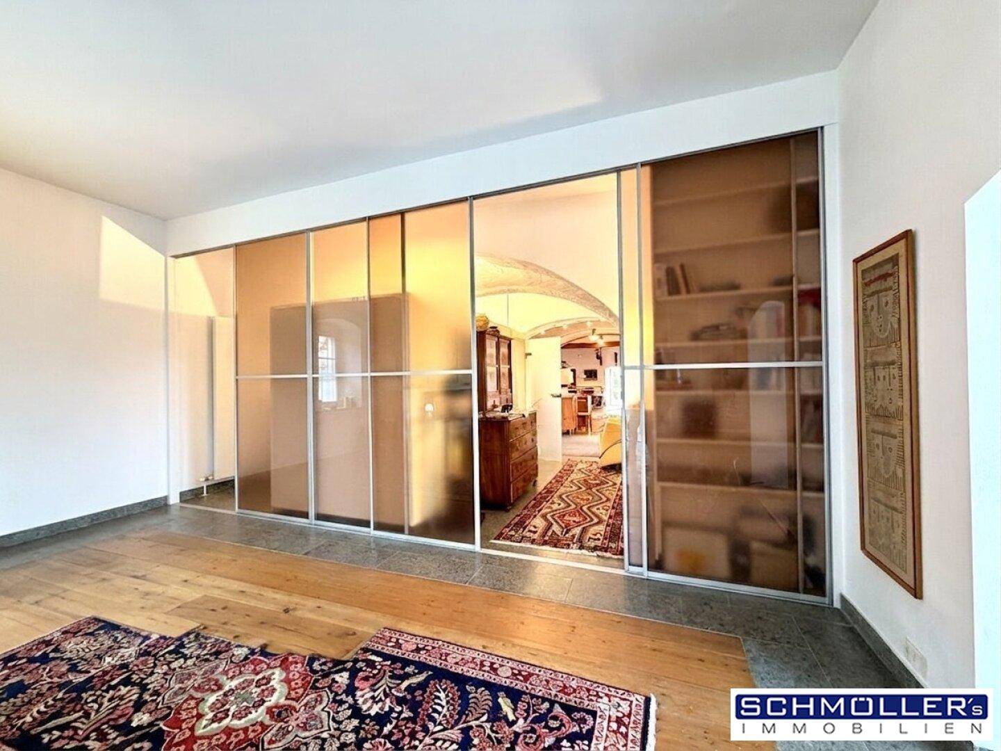 Heller Wohnbereich mit Holzboden, großem Teppich und Blick durch eine Glaswand in den angrenzenden Raum.