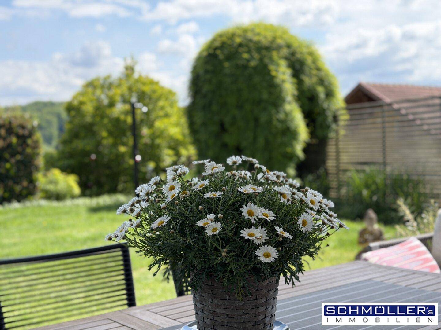 Sonnige Terrasse mit Gartenblick und Blumenarrangement auf einem Holztisch.