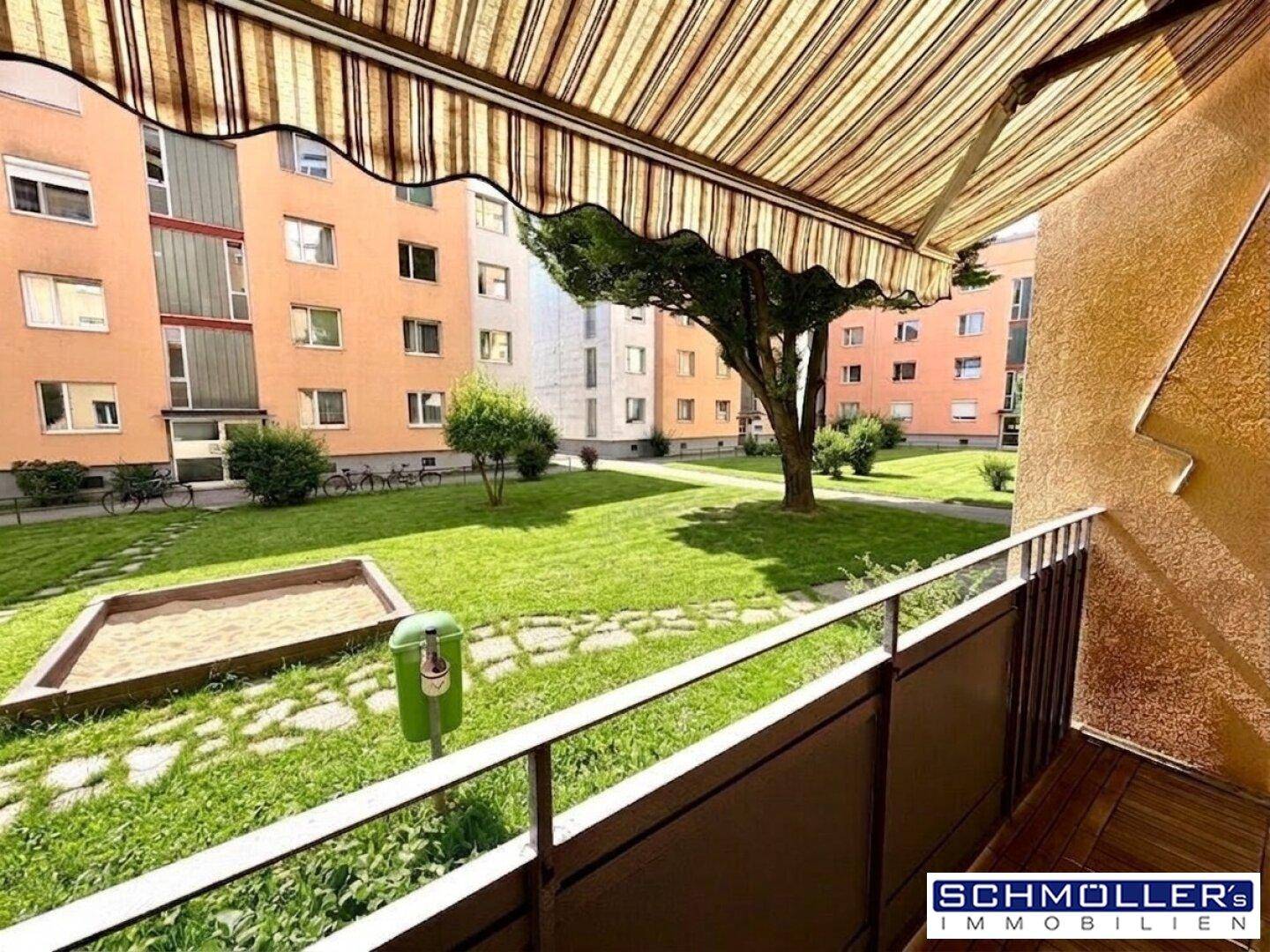 Blick vom Balkon mit gestreifter Markise auf einen grünen Innenhof mit Sandkasten und umliegenden Wohngebäuden.