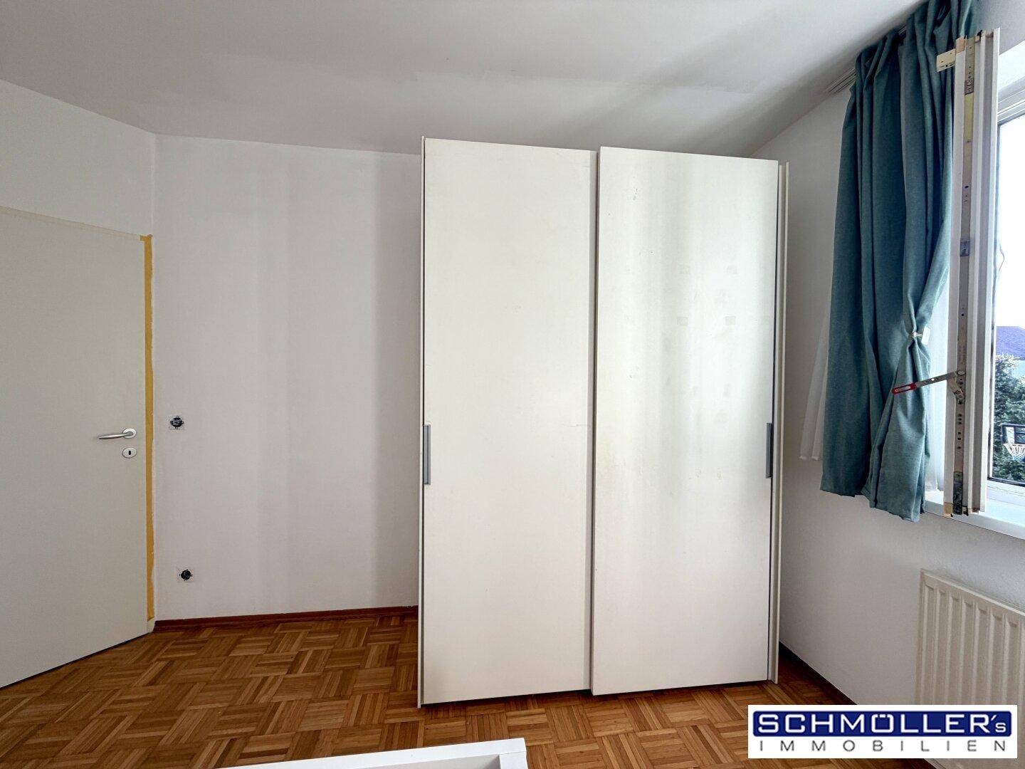 Schlafzimmer mit Schiebetürenschrank, Fenster und Parkettboden.