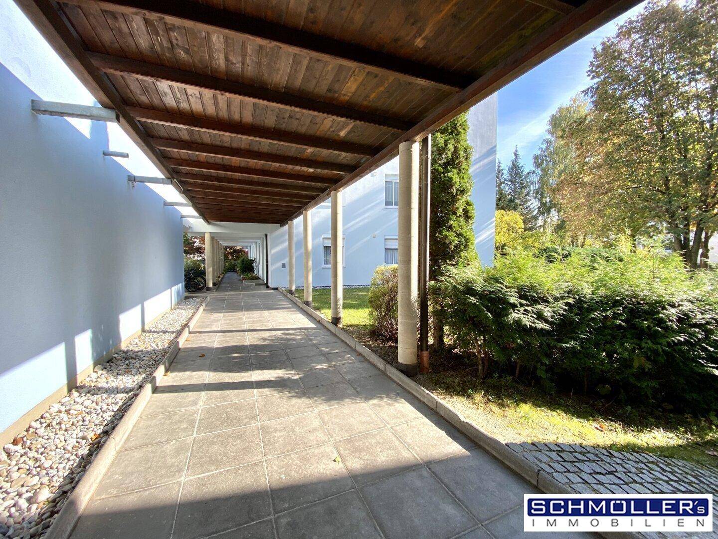Überdachter Gehweg mit Holzdach und Kiesbeet entlang eines Wohngebäudes an einem sonnigen Tag.