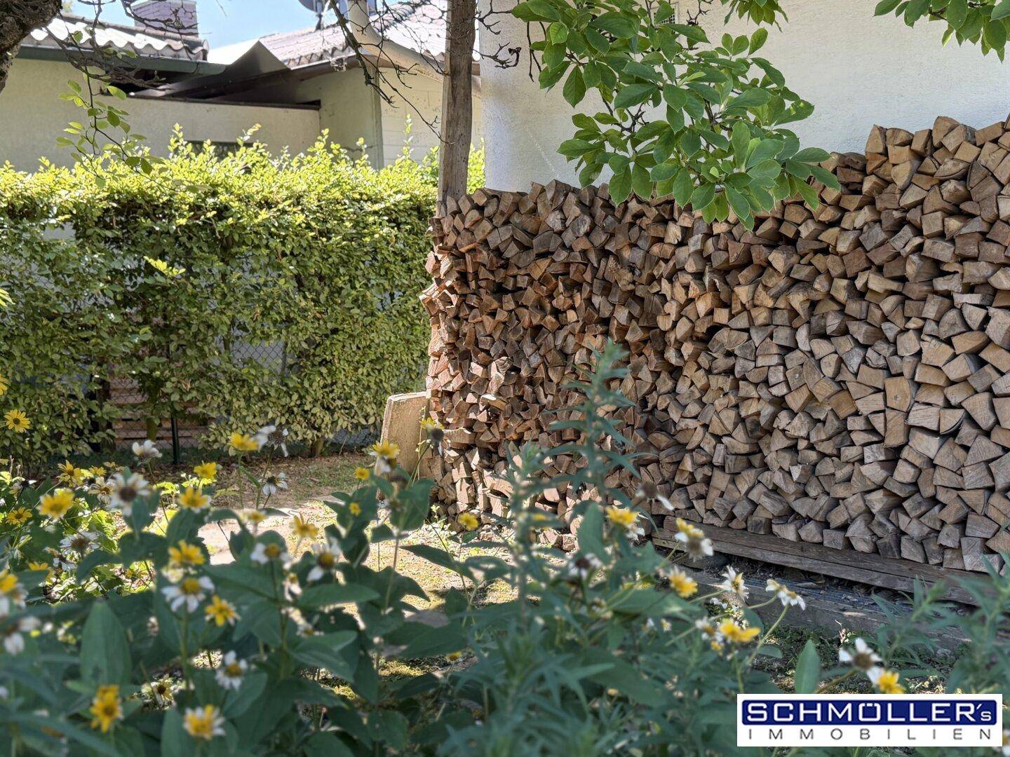 Großer Stapel Brennholz im Garten, umgeben von blühenden Pflanzen und einer Hecke.