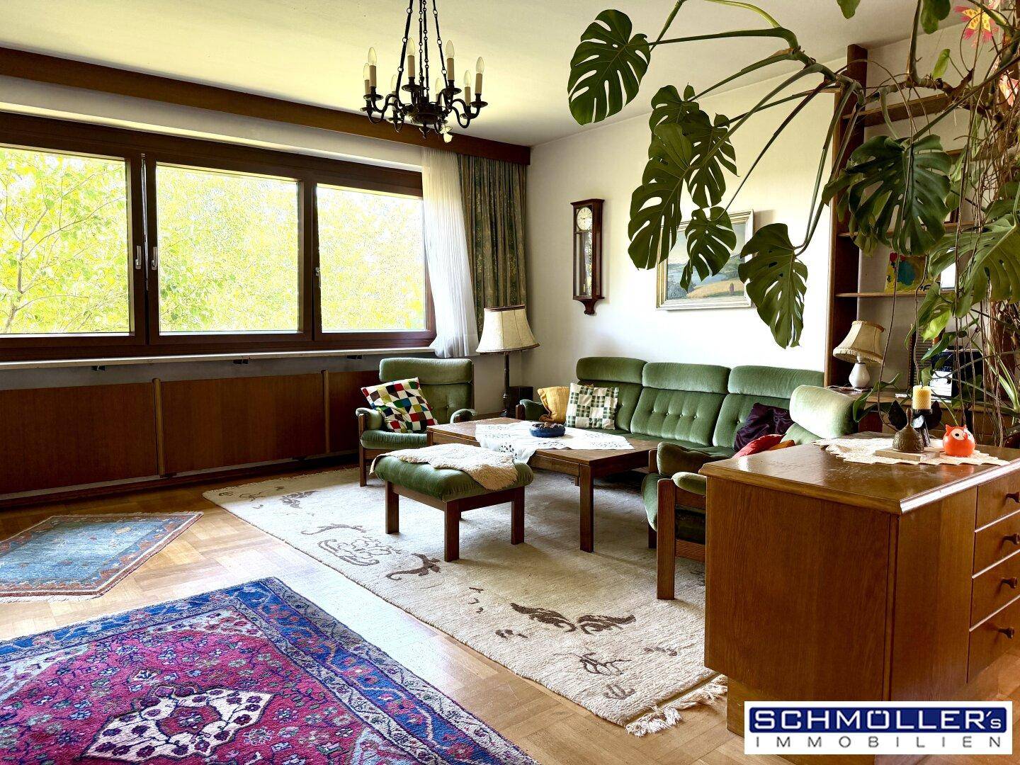 Geräumiges Wohnzimmer mit grünen Sofas, Holzboden und großen Fenstern mit Blick ins Grüne.