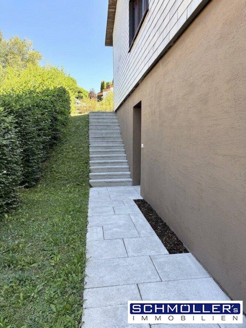 Gepflasterter Weg und Treppe führen entlang einer Hecke zum Eingang des Hauses.
