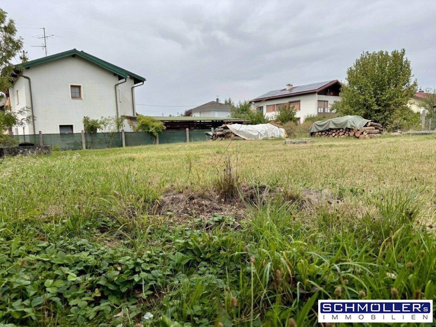 Weitläufiges Grundstück mit hohem Gras und Holzstapeln im Hintergrund, umgeben von Wohnbebauung.