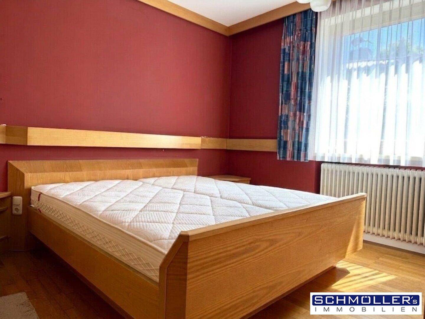 Schlafzimmer mit roter Wand, Holzbett, Parkettboden und einem Fenster mit Heizkörper darunter.