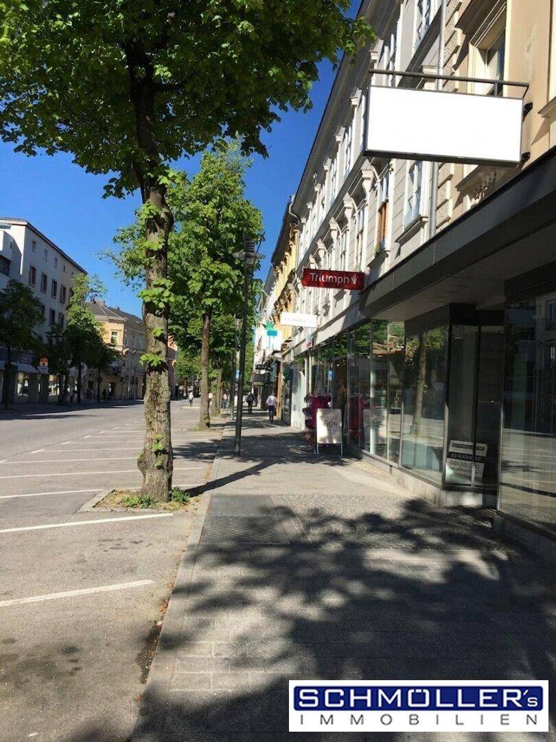Belebtes Stadtbild mit Geschäften, Bäumen und parkenden Autos entlang einer breiten Straße.