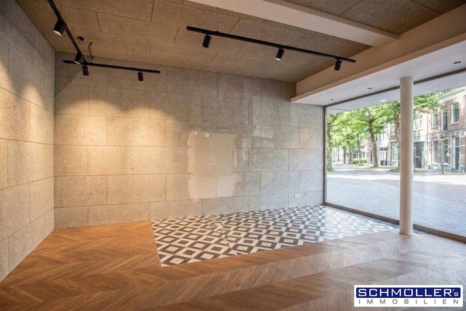 Heller Geschäftsraum mit großen Fenstern, Holzboden und strukturierten Wänden, ideal für Präsentationen.