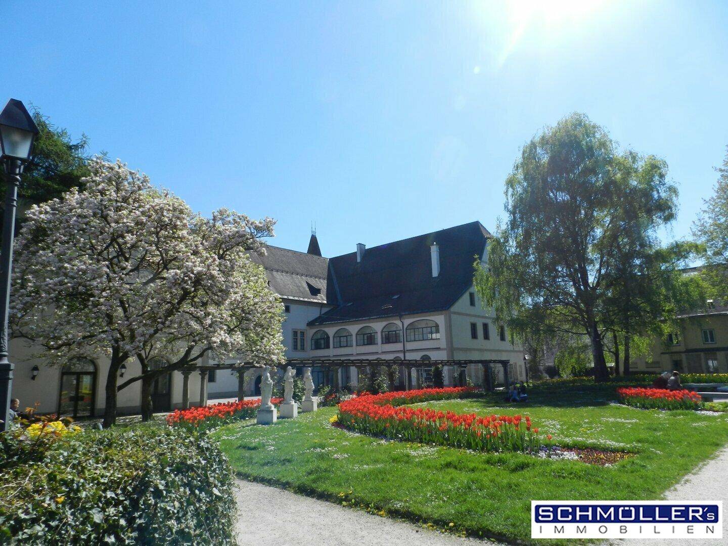 Gepflegter Gartenbereich mit blühenden Bäumen, roten Tulpen und einem historischen Gebäude im Hintergrund.