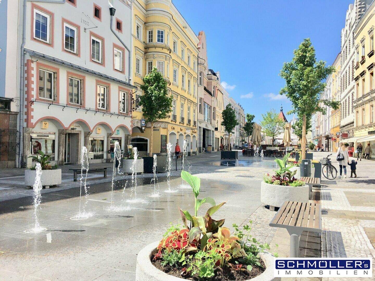 Malerischer Stadtplatz mit Springbrunnen, Blumenbeeten und historischen Gebäuden unter blauem Himmel.