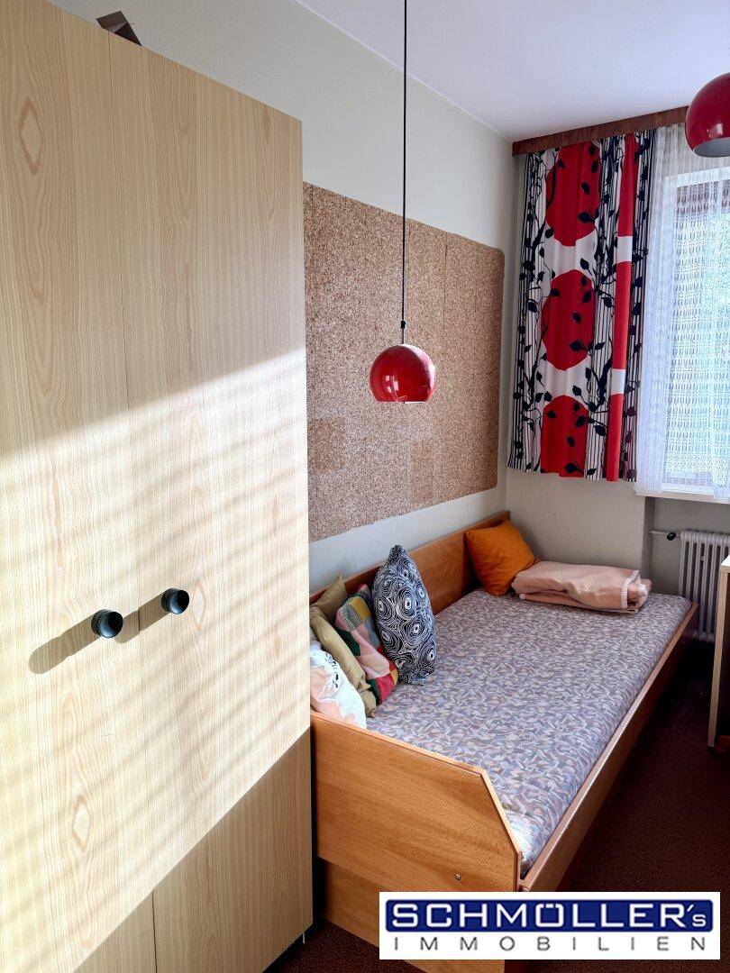 Helles Schlafzimmer mit Einzelbett, hellem Holzschrank und farbenfrohen Vorhängen am Fenster.
