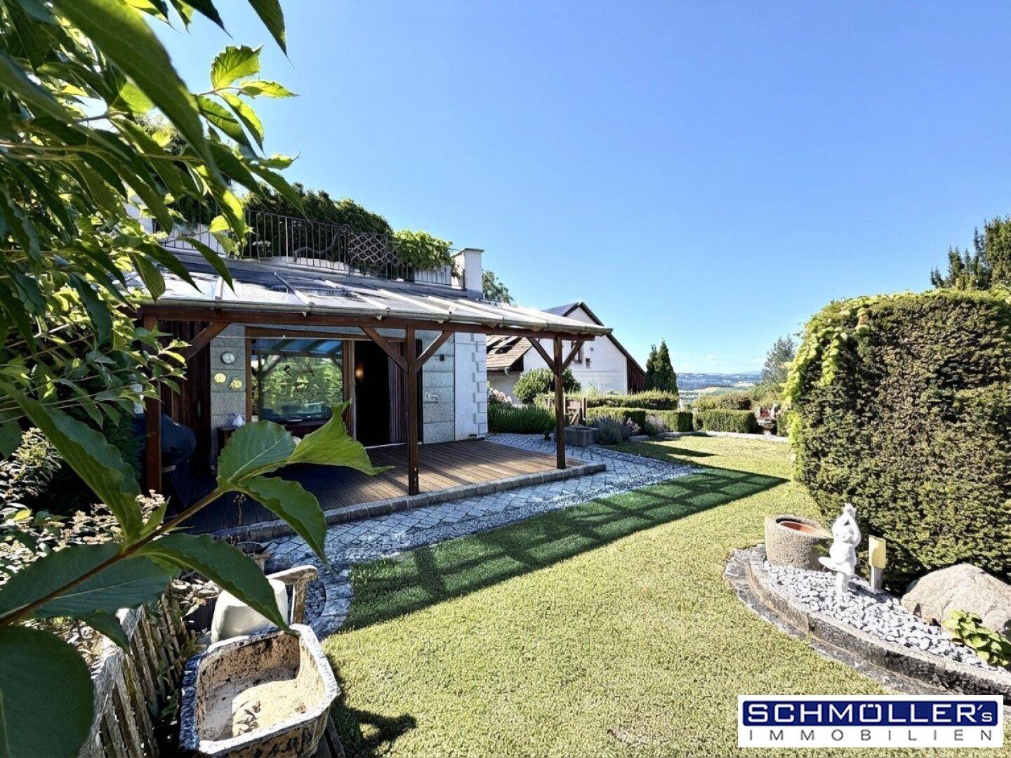 Sonniger Garten mit Rasenfläche, Sträuchern und einer Holzterrasse im Hintergrund unter klarem Himmel.