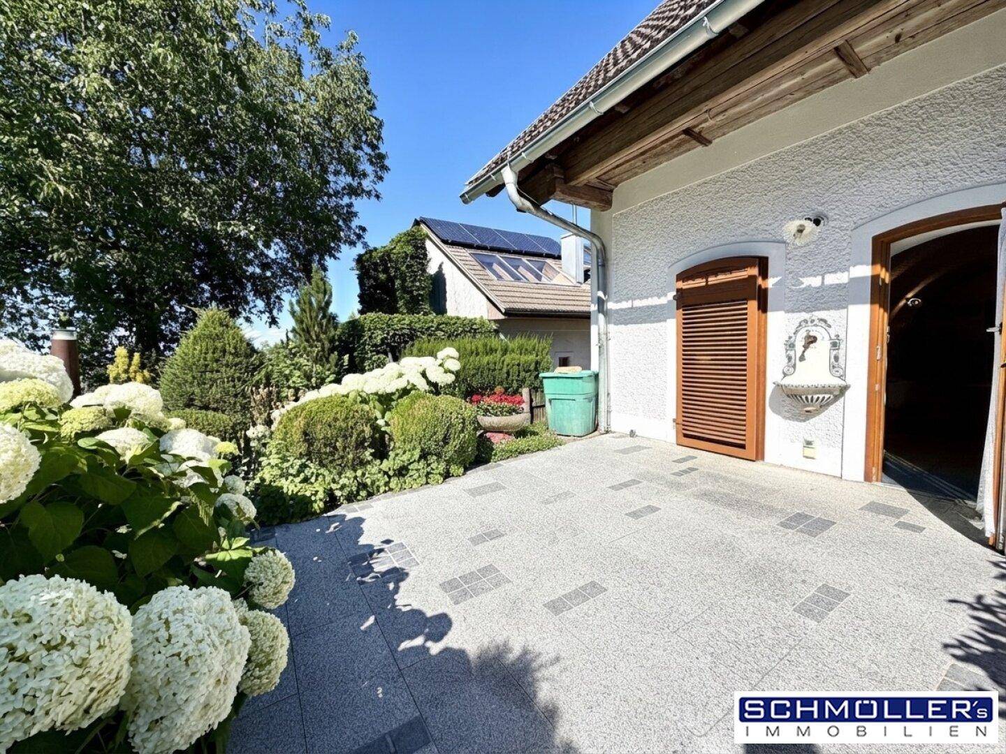 Gepflasterte Terrasse mit blühenden Hortensien und Blick auf ein Haus mit Solaranlage im Hintergrund.