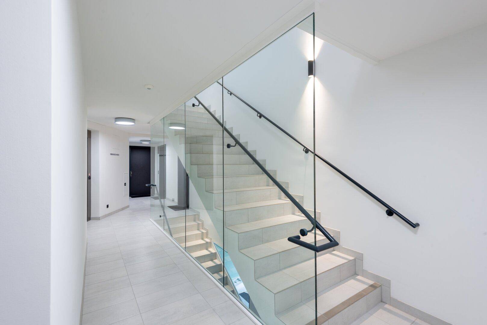 Heller, moderner Flur mit eleganter Treppe, Glasgeländer und schwarzen Handläufen.