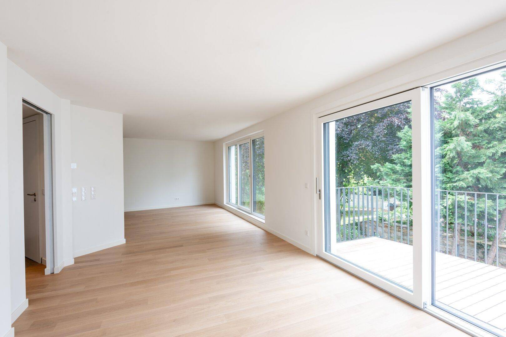 Helles, leeres Wohnzimmer mit Eichenparkett und Zugang zu einem Balkon mit Blick ins Grüne.