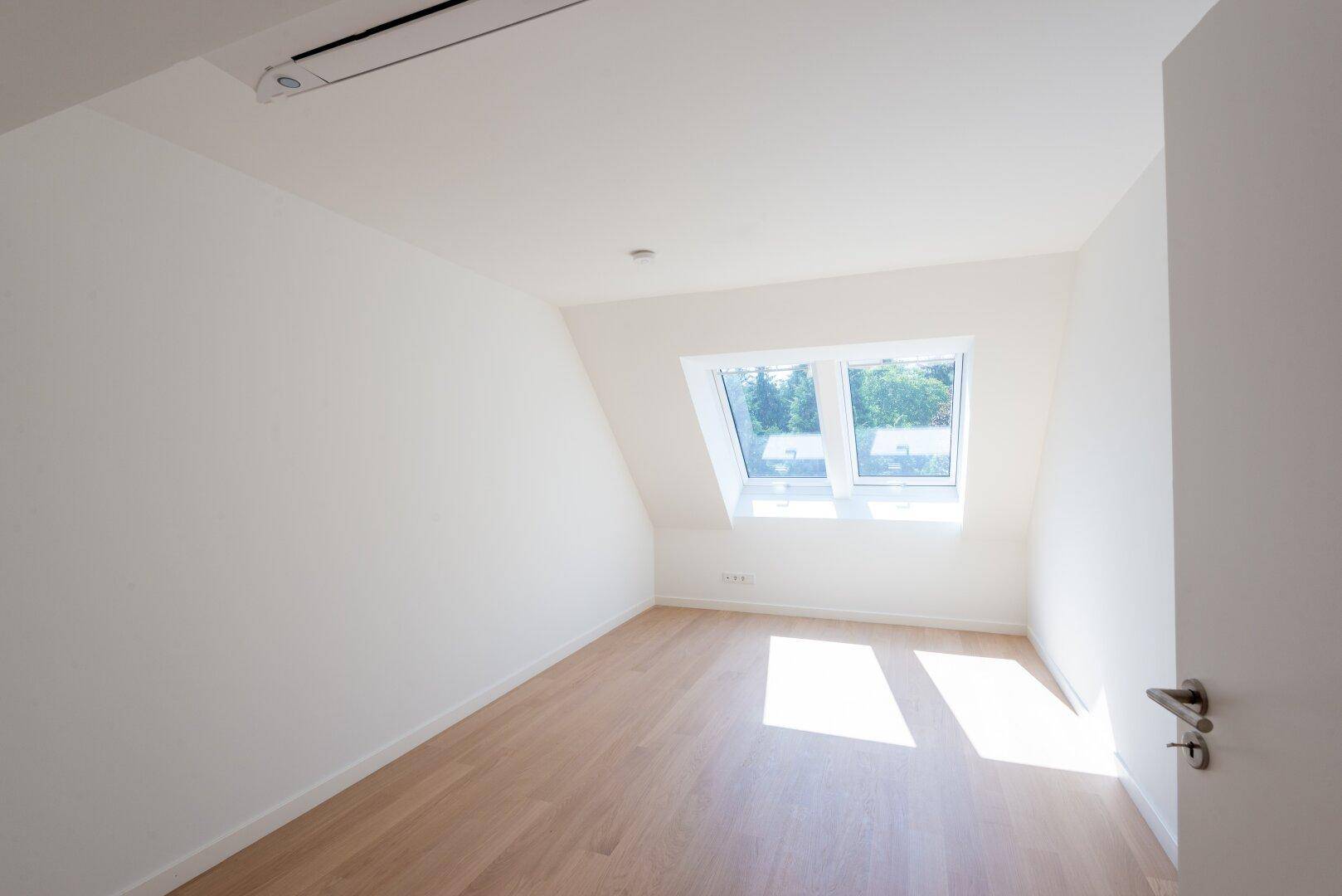 Helles Zimmer im Dachgeschoss mit schrägen Wänden, Holzboden und Dachfenstern für viel Tageslicht.
