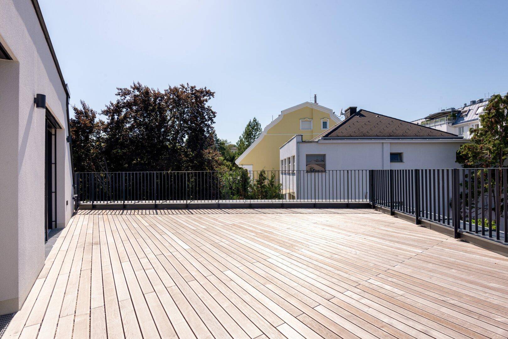 Große Dachterrasse mit hellem Holzbelag und Blick auf umliegende Gebäude und grüne Bäume.