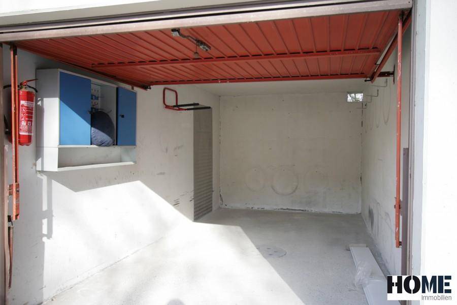 Leere Garage mit rotem Rolltor, Betonboden und einem Feuerlöscher an der Wand.