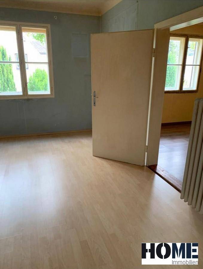 Leerer Raum mit hellblau-grauen Wänden und Laminatboden, angrenzend an einen weiteren Raum mit gelben Wänden.
