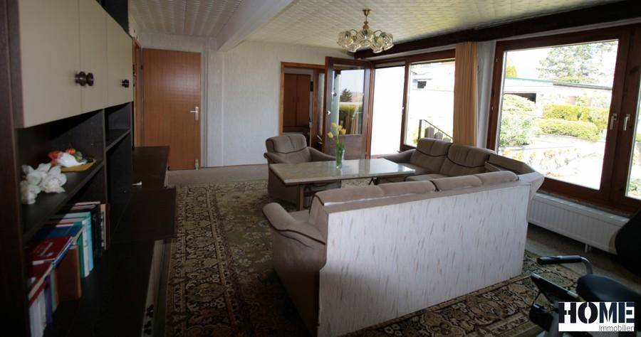 Geräumiges Wohnzimmer mit bequemer Sitzecke, großen Fenstern und Blick ins Grüne.