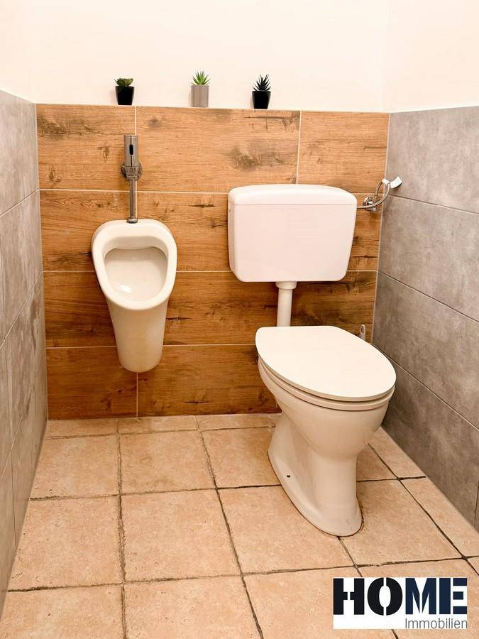 Herren-WC mit Toilette und Urinal, ausgestattet mit Fliesenboden und Holzverkleidung an der Wand.