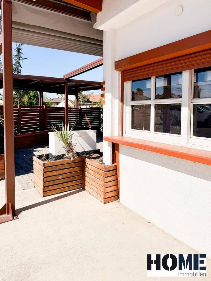 Außenansicht des Gebäudes mit Fenster, Holzrollläden und dekorativen Pflanzenkübeln auf gepflastertem Boden.