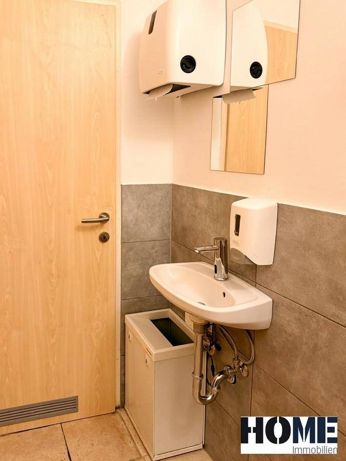 Toilettenraum mit Waschbecken, Spiegel und Spender für Papierhandtücher, in neutralen Farben gehalten.