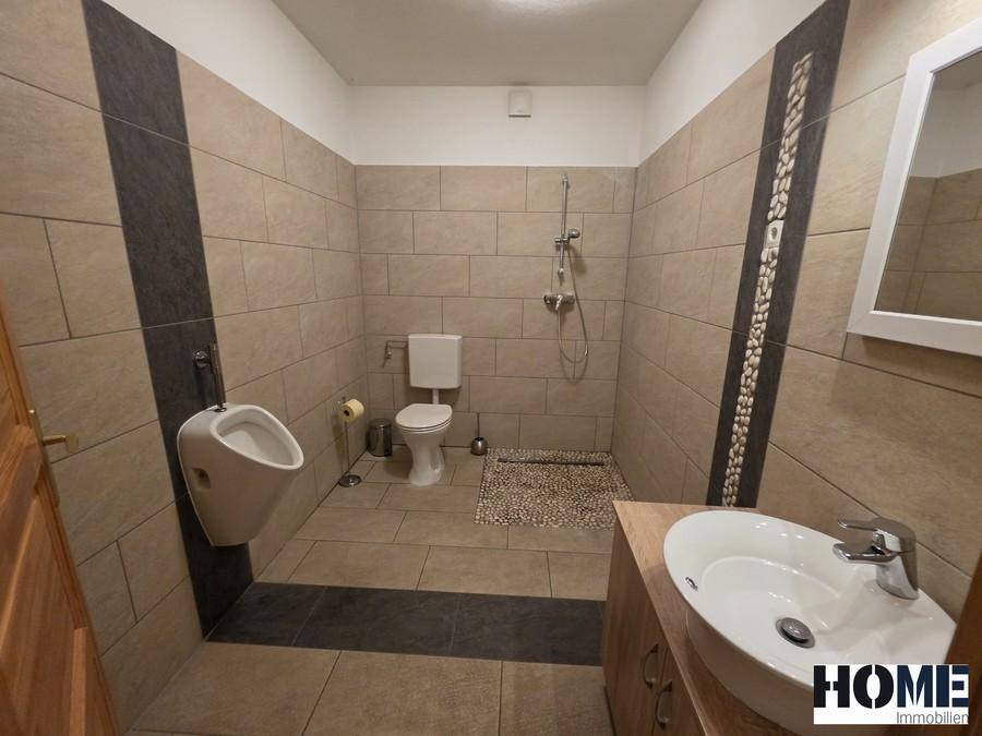 Modernes Badezimmer mit beigefarbenen Fliesen, Dusche, Urinal und Einzelwaschbecken.