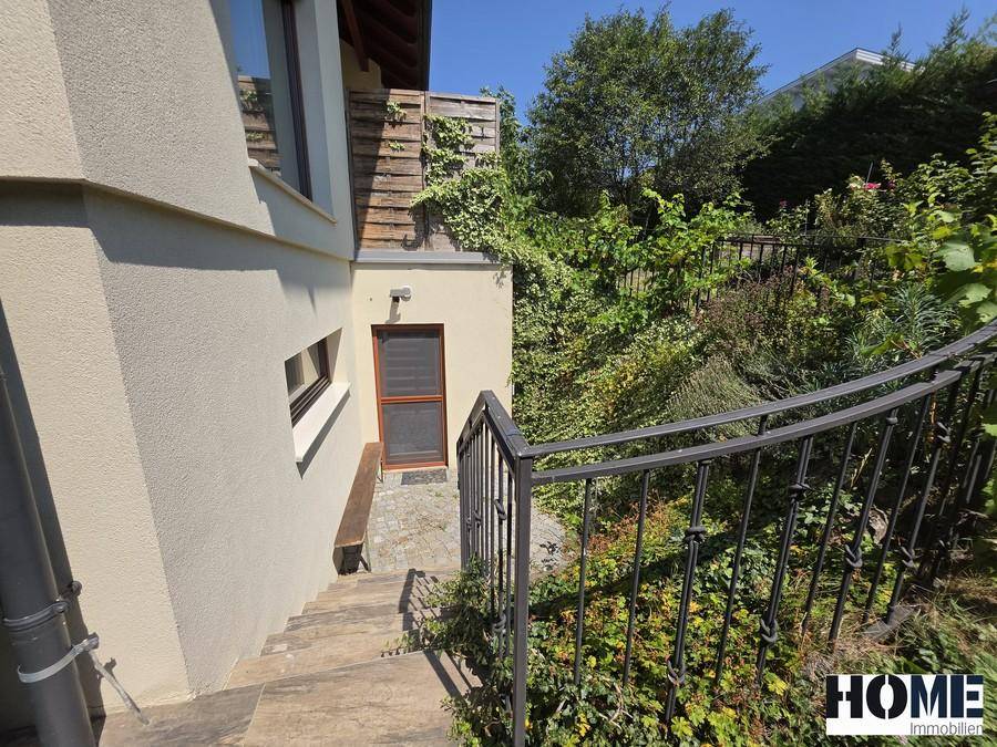 Außentreppe führt hinunter zu einem Eingang, umgeben von üppigem Grün und einer Metallbalustrade.