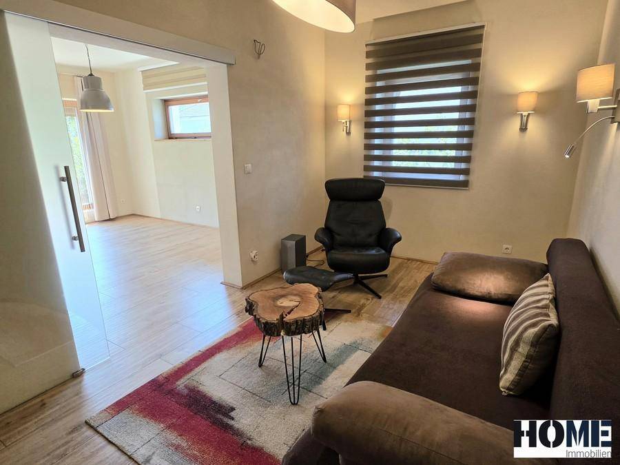 Helles Wohnzimmer mit Schiebetür, Sofa und Sessel, verbunden mit einem weiteren Wohnbereich.
