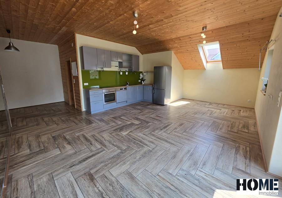 Offener Wohn- und Küchenbereich mit schräger Holzdecke, Holzbodenoptik und einer modernen grauen Küche.