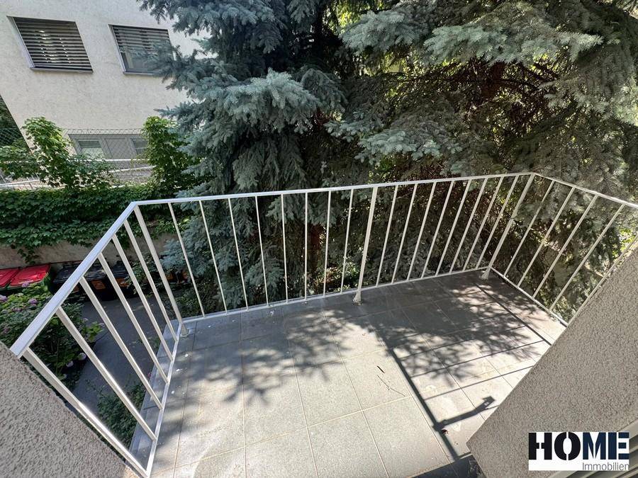 Kleiner Balkon mit Fliesenboden und weißem Geländer, umgeben von üppigem Grün und Bäumen.