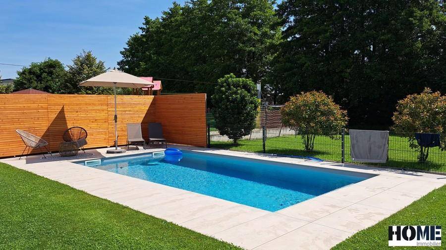 Sonniger Poolbereich mit Liegewiese, Holzumzäunung und umgebenden Bäumen.