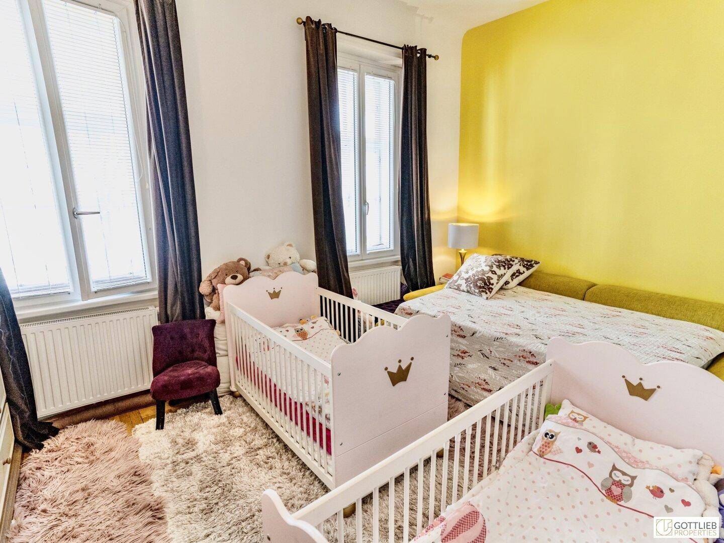 Helles Kinderzimmer mit zwei Babybetten, einem Einzelbett und einer freundlichen gelben Wand.