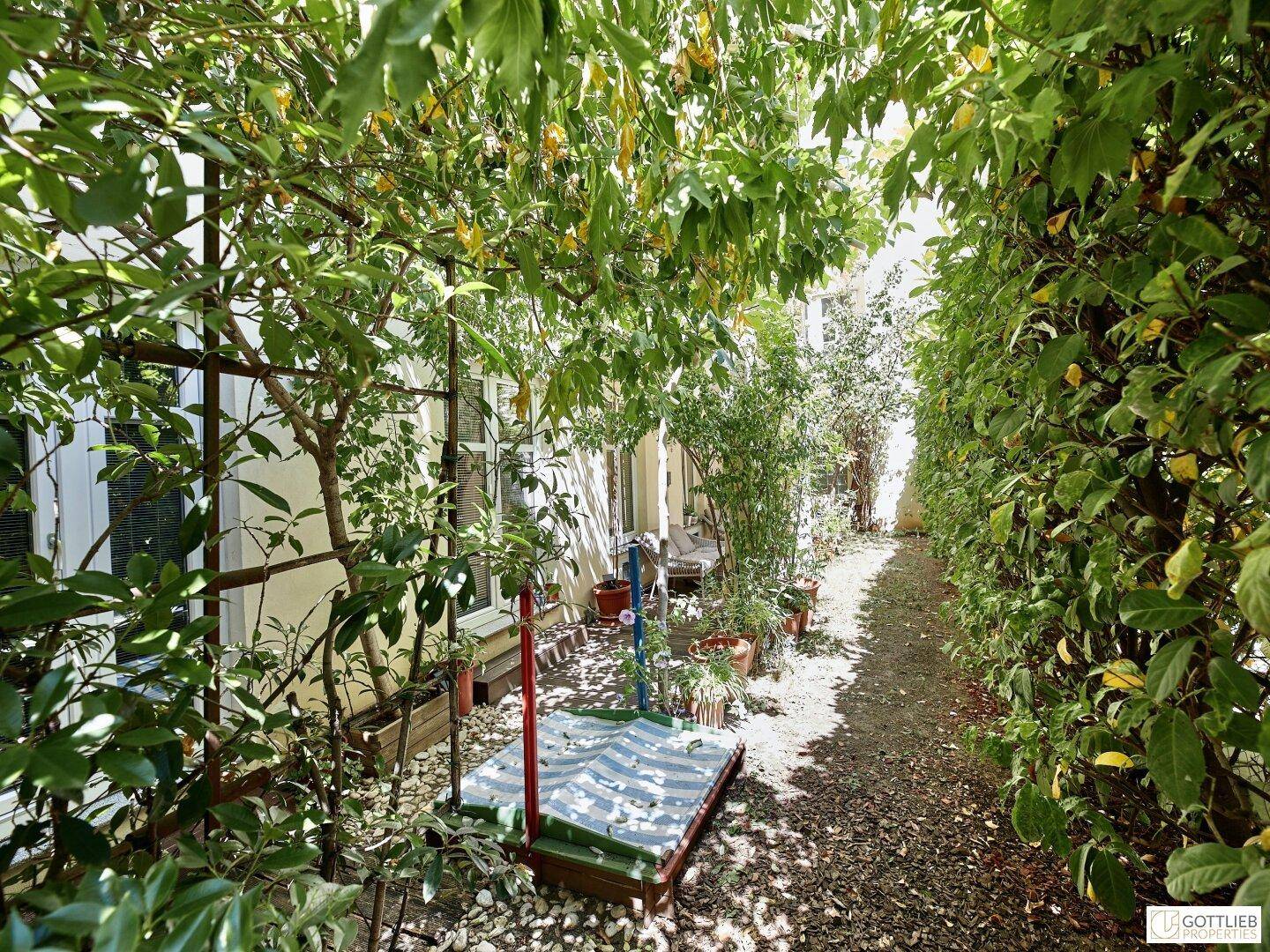 Schmaler Gartenweg umgeben von dichtem Grün, der zu einem kleinen Sitzbereich führt.
