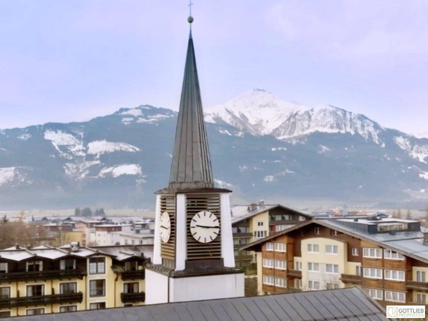 Kirchturm mit Uhr vor einer Bergkulisse und umliegenden Gebäuden.