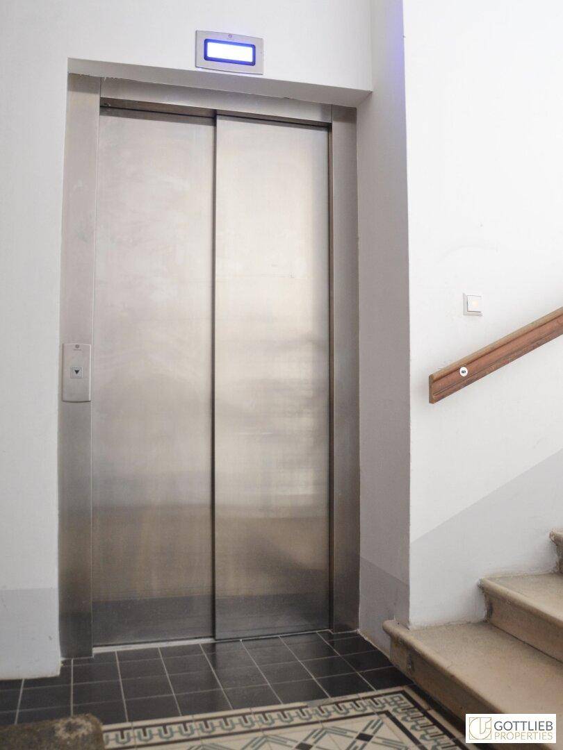 Moderner Aufzug in einem Treppenhaus mit Fliesenboden und Holzgeländer.