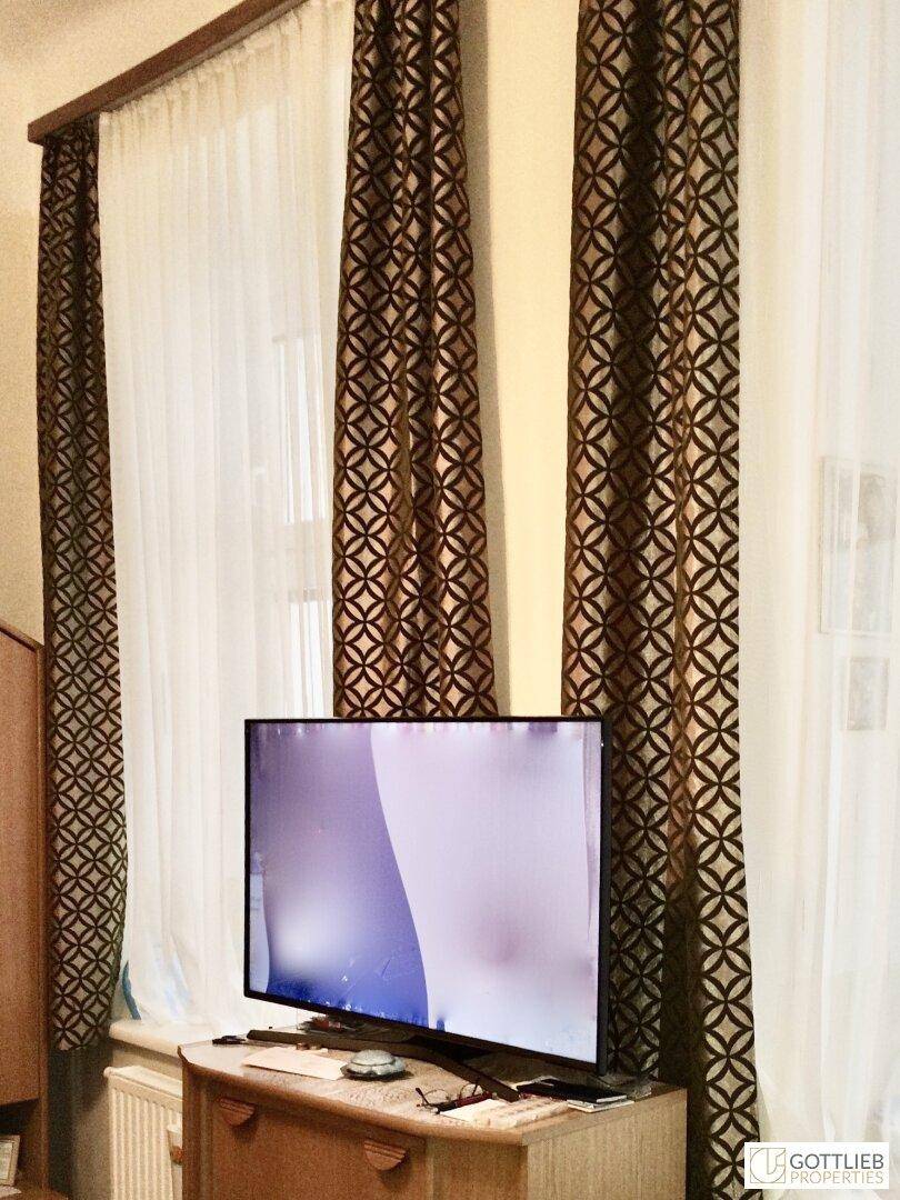 Fernseher auf einem Holzschrank vor einem Fenster mit gemusterten Vorhängen in einem Wohnbereich.