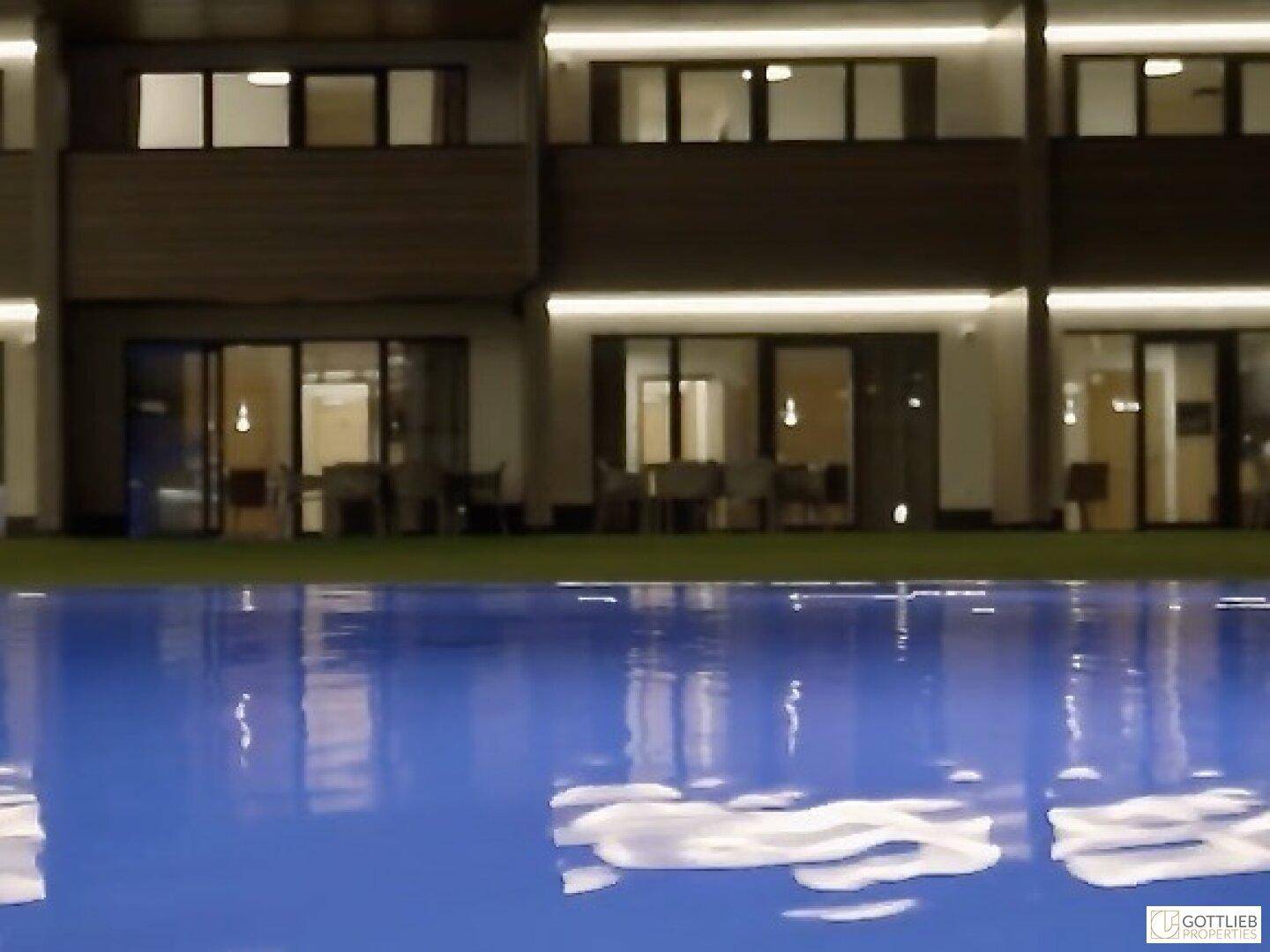 Beleuchteter Außenpool vor einem modernen Gebäude mit hellen Balkonen und Terrassen bei Nacht.