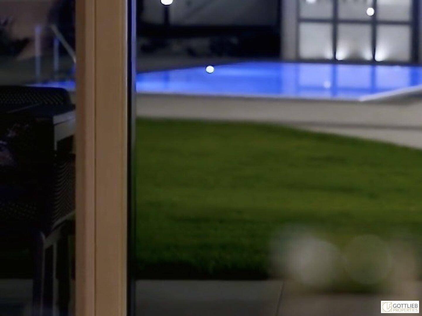 Unscharfer Blick aus dem Fenster auf einen beleuchteten Pool und eine Rasenfläche bei Nacht.