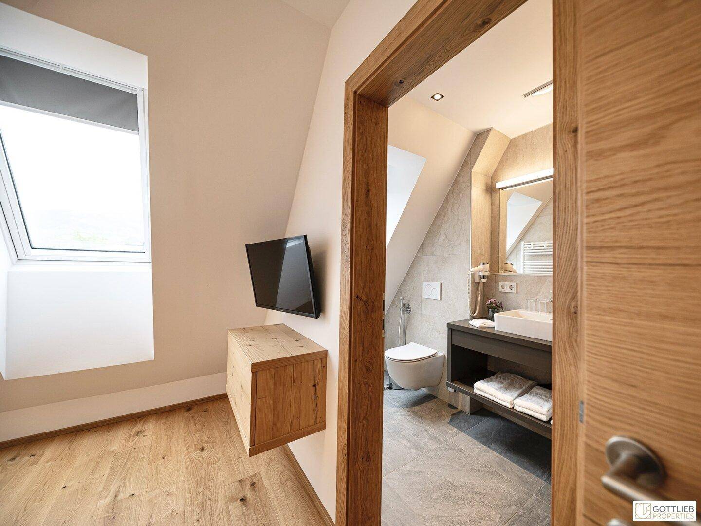 Schlafzimmer im Dachgeschoss mit Holzboden, Fernseher und Blick auf das angrenzende Badezimmer.