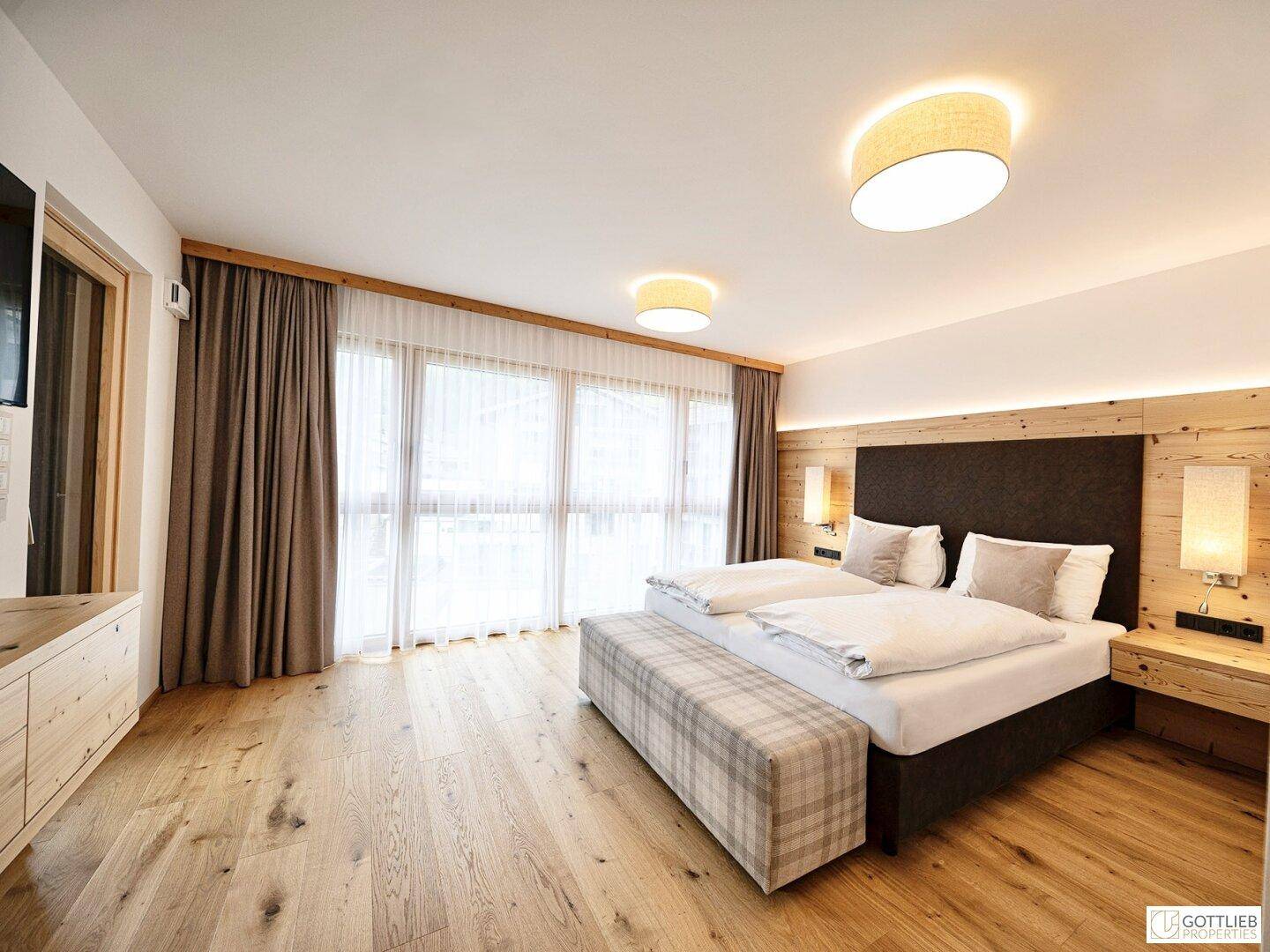 Helles Schlafzimmer mit Doppelbett, Holzboden und großen Fenstern mit Vorhängen.