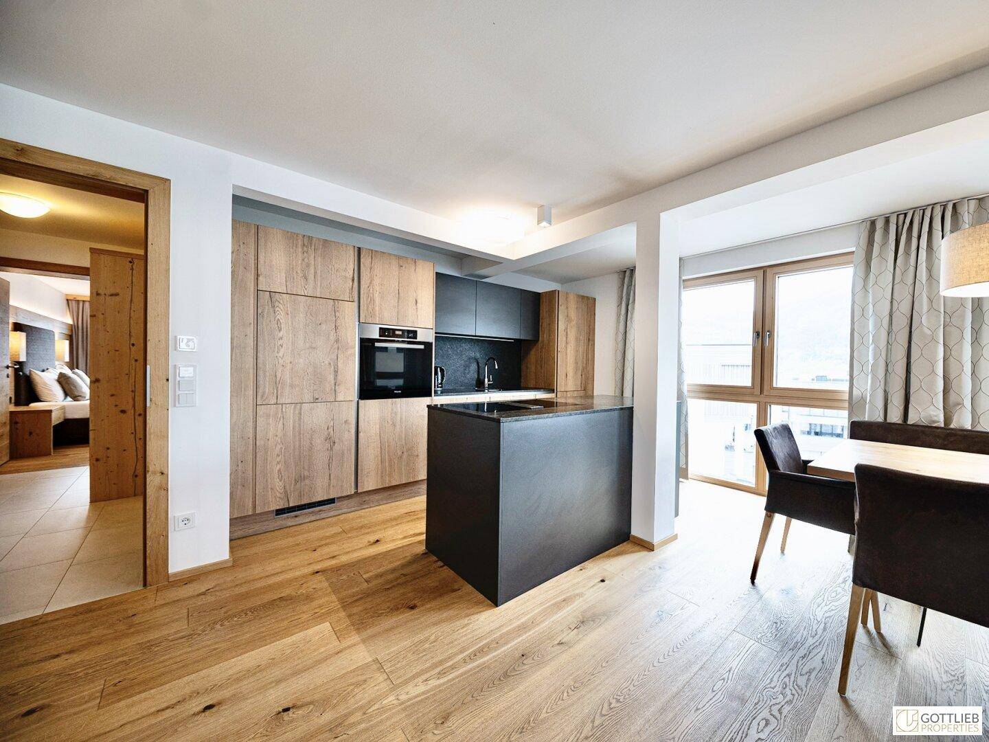 Offener Wohnbereich mit moderner Küche, Essplatz und Holzboden, der zu einem Schlafzimmer führt.