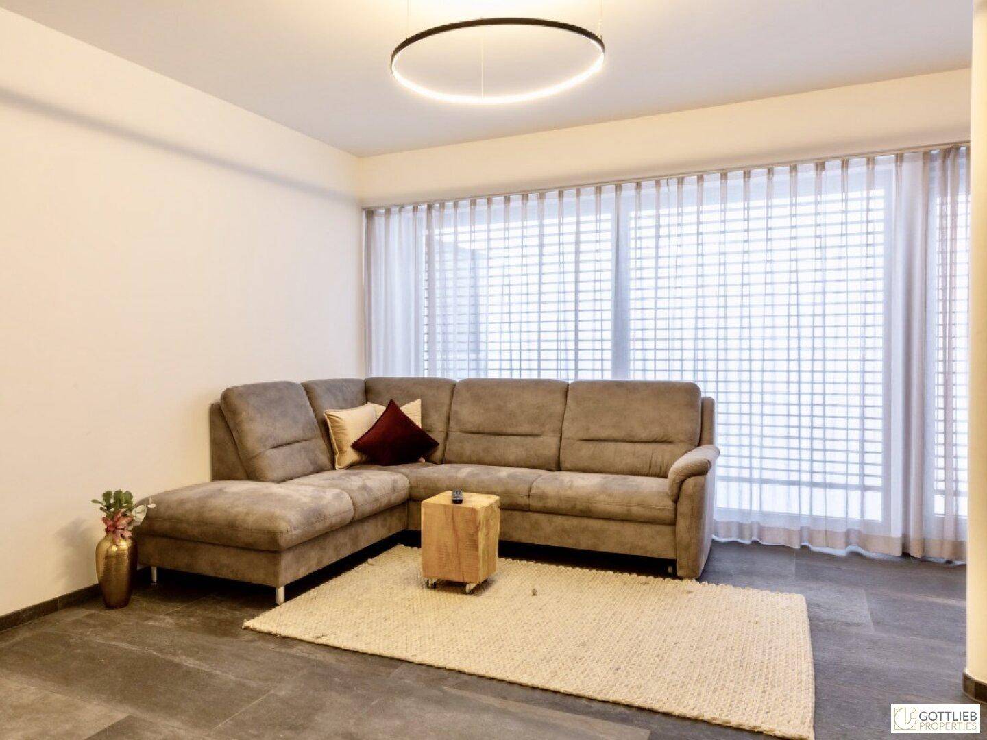 Gemütliches Wohnzimmer mit großer Eckcouch, Teppich und bodentiefen Fenstern.