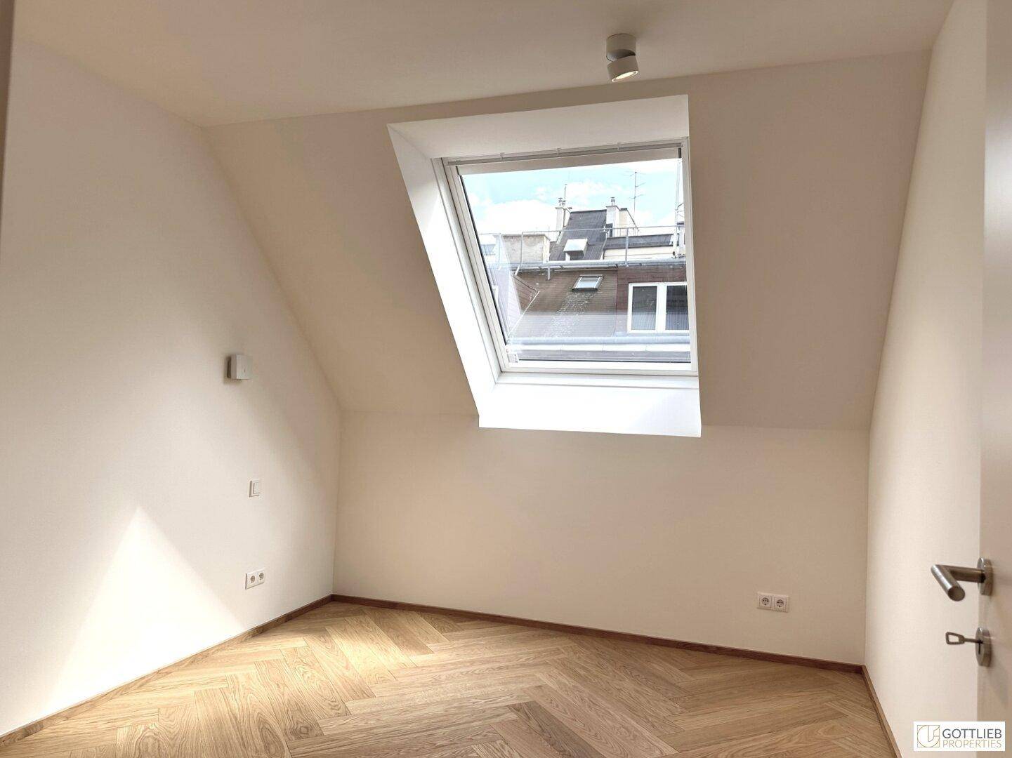 Ruhiges Zimmer im Dachgeschoss mit schrägen Wänden und einem großen Dachfenster für viel Licht.