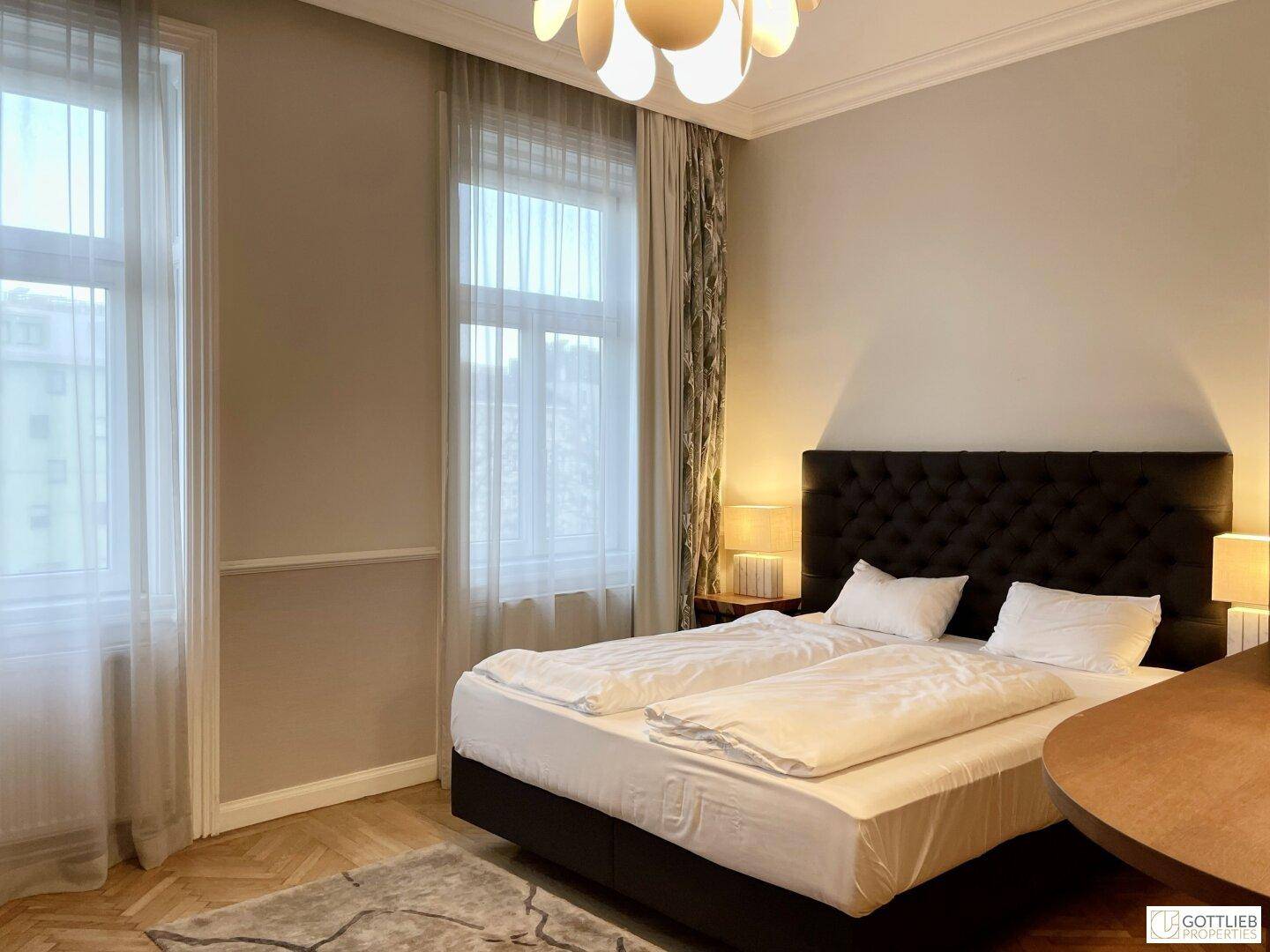Gemütliches Schlafzimmer mit großem Doppelbett, zwei hohen Fenstern und eleganter Deckenleuchte.