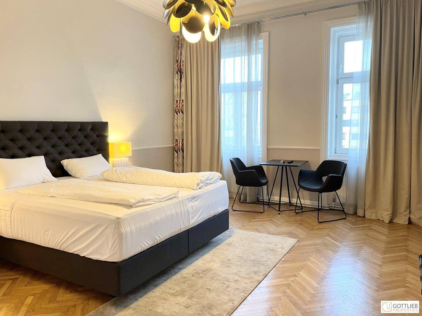 Geräumiges Studio-Apartment mit Doppelbett, großen Fenstern und einem kleinen Tisch mit Stühlen.