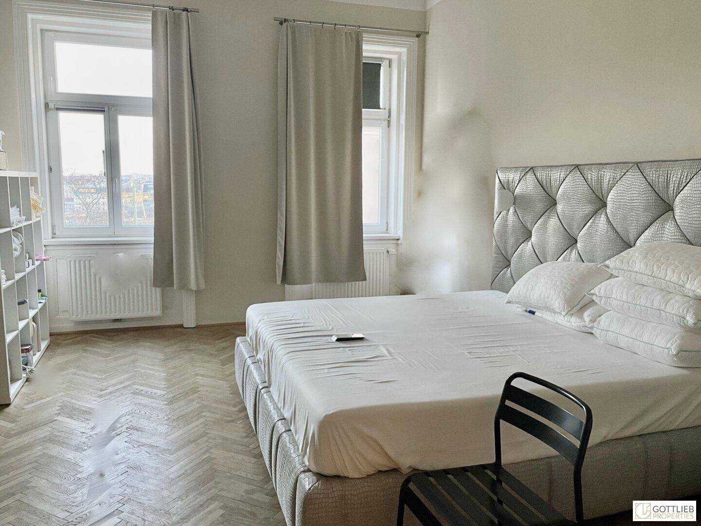 Geräumiges Schlafzimmer mit großem Bett, zwei Fenstern und einem praktischen Regal für Bücher oder Dekoration.