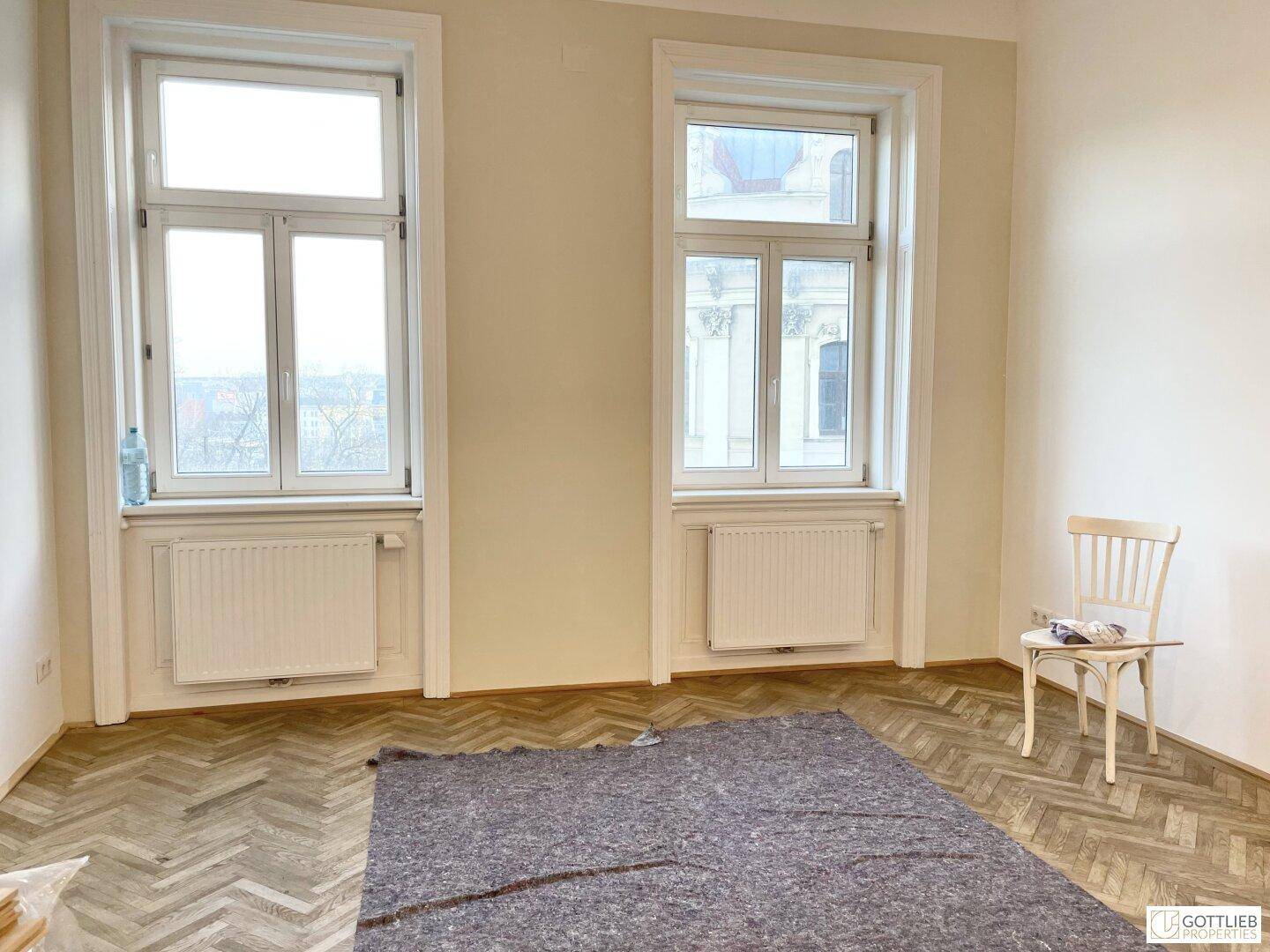 Heller, leerer Raum mit Fischgrätparkett, zwei Fenstern und einem Teppich, bereit zur Einrichtung.