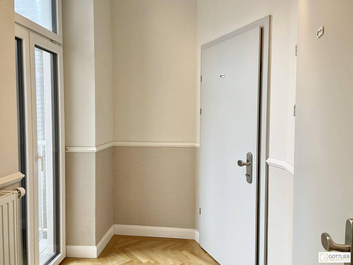 Eingangsbereich mit Fischgrätparkett, einer Tür und einem Fenster, das Zugang zu einem Balkon bietet.